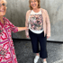 Zwei lachende Frauen in modischer Kleidung, eine trägt eine rosa Jacke und ein T-Shirt mit Print vor einer grauen Wand.