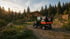 Kleiner schwarzer Elektrolieferwagen mit orange-schwarzem Logo auf Schotterweg vor Berghütte und Wald bei Sonnenuntergang.