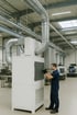 Mann in blauer Arbeitskleidung justiert eine weiße Industriemaschine mit großen Metallrohren in einer Fabrikhalle.