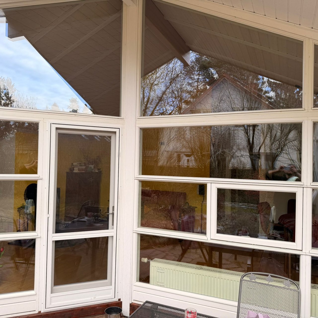 Ein Wintergarten mit vielen Fenstern und einer Tür; draußen sind Bäume und ein Haus zu sehen.