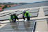 Zwei Arbeiter in Schutzkleidung installieren Solarmodule auf einem wellblechgedeckten Dach.