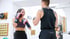 Eine lächelnde Frau mit Boxhandschuhen trainiert mit einem Partner in einem hellen Boxclub.