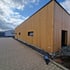 Modernes Holzgebäude im Bau mit brauner Holzverkleidung, schwarzen Fenstern und gepflastertem Gehweg.