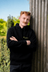 Ein Mann lehnt in einem schwarzen Hoodie an einer Holzwand im Garten.
