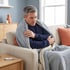 A man wrapped in a grey blanket sits on a sofa next to a digital thermometer on a wooden side table.