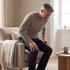 A man with gray hair winces while holding his knee as he attempts to stand up from a beige armchair.