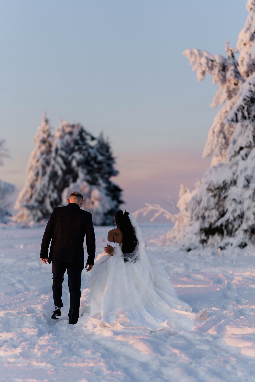 Brautpaar geht Hand in Hand durch den Schnee, umgeben von verschneiten Tannen im Abendlicht in Winterberg.