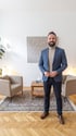 Ein lächelnder Mann mit Bart, blauem Blazer und beigem Rollkragenpullover steht in einem Therapieraum.