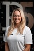 Lächelnde blonde Physiotherapeutin im weißen Adidas-Shirt mit Logo der Praxis Therapier und Pier5  - this is PHYSIO vor dunklem Hintergrund.