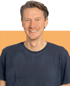 Ein lächelnder Mann mit braunen Haaren und einem dunkelblauen T-Shirt vor einem orange-grünen Hintergrund.