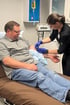 A nurse in black scrubs inserts an IV into the arm of a man sitting on a medical exam table.