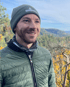 Ein Mann mit Mütze, Jacke und Halstuch lächelt in einer Berglandschaft mit Herbstbäumen.
