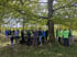 Eine Gruppe von Wanderern rastet an einem Picknicktisch im Schatten großer, grüner Laubbäume auf einer Waldlichtung.