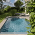 Rechteckiger Pool mit bläulichem Wasser in einem Garten, umgeben von grünen Pflanzen, Rasen und einer Terrasse.