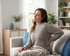 A woman sits on a sofa with a pained expression, holding her lower back in a living room setting.