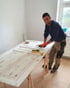 Handwerker bearbeitet eine alte weiße Holztür mit einem Spachtel auf zwei Holzböcken in einem hellen Raum.