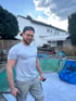 Simon Tomeczek, Poolbau-Profi in der Rhein-Ruhr Region (NRW) mit Kappe und Brille, der einen Hammer und ein Metallstück hält, steht vor einem leeren Pool und einem Haus.