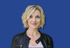 Eine lächelnde Frau mit blonden Haaren und schwarzer Lederjacke vor blauem Hintergrund.