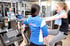 Zwei Frauen unterhalten sich im Fitnessstudio, eine davon trägt ein blaues T-Shirt mit der Aufschrift „fit team“.