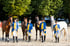 Vier Reiter in blau-gelben Uniformen stehen mit ihren Pferden auf einem Reitplatz vor einer bewaldeten Kulisse.