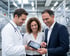 Ein Arzt, eine Frau und ein Geschäftsmann betrachten lächelnd ein Tablet in einer modernen Industrieanlage.