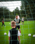 Ein Fußballtornetz im Vordergrund, dahinter ein Kind, das einem Fußballtrainer auf einem Rasenplatz zuhört.