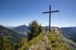Gipfelkreuz auf einem Felsen mit grüner Wiese und Bäumen, umgeben von einem weiten Tal und Bergen unter blauem Himmel.