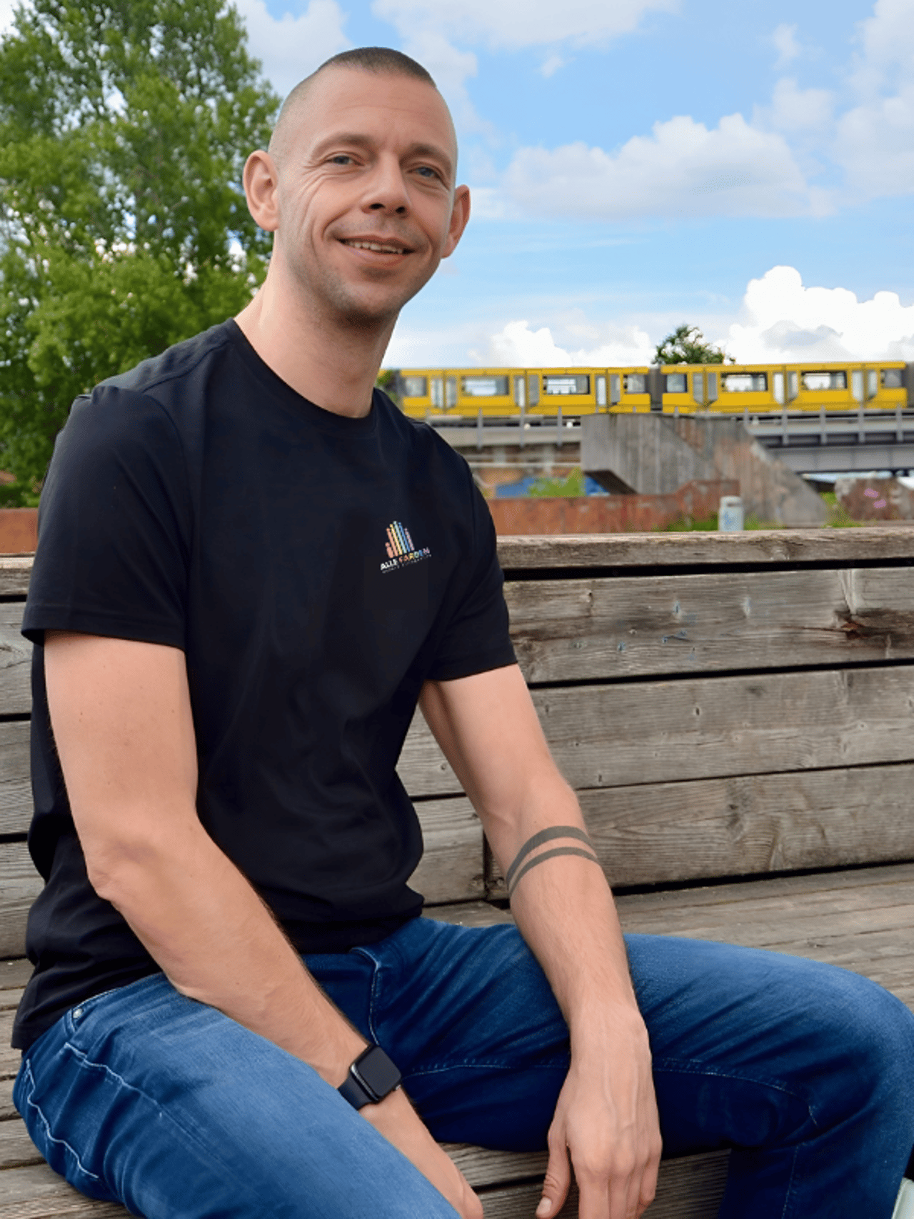 Mann in schwarzem T-Shirt und Jeans sitzt lächelnd auf Holzbank; gelbe U-Bahn fährt im Hintergrund.