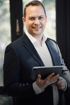 Ein Mann in einem Anzug hält ein Tablet und lächelt in die Kamera.