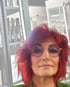 Frau mit kurzen roten Haaren und runder Brille blickt in die Kamera, Hintergrund ist ein Friseursalon.