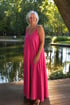 Lächelnde Frau mit grauen Haaren in einem langen pinken Kleid steht auf einem Holzsteg vor einem See im Park.