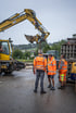 Drei Bauarbeiter in orangefarbenen Warnwesten beobachten einen gelben Liebherr Bagger mit Schaufel.