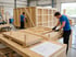 Zwei Handwerker in blauen T-Shirts fertigen große Transportkisten aus Holz in einer Industriehalle an.