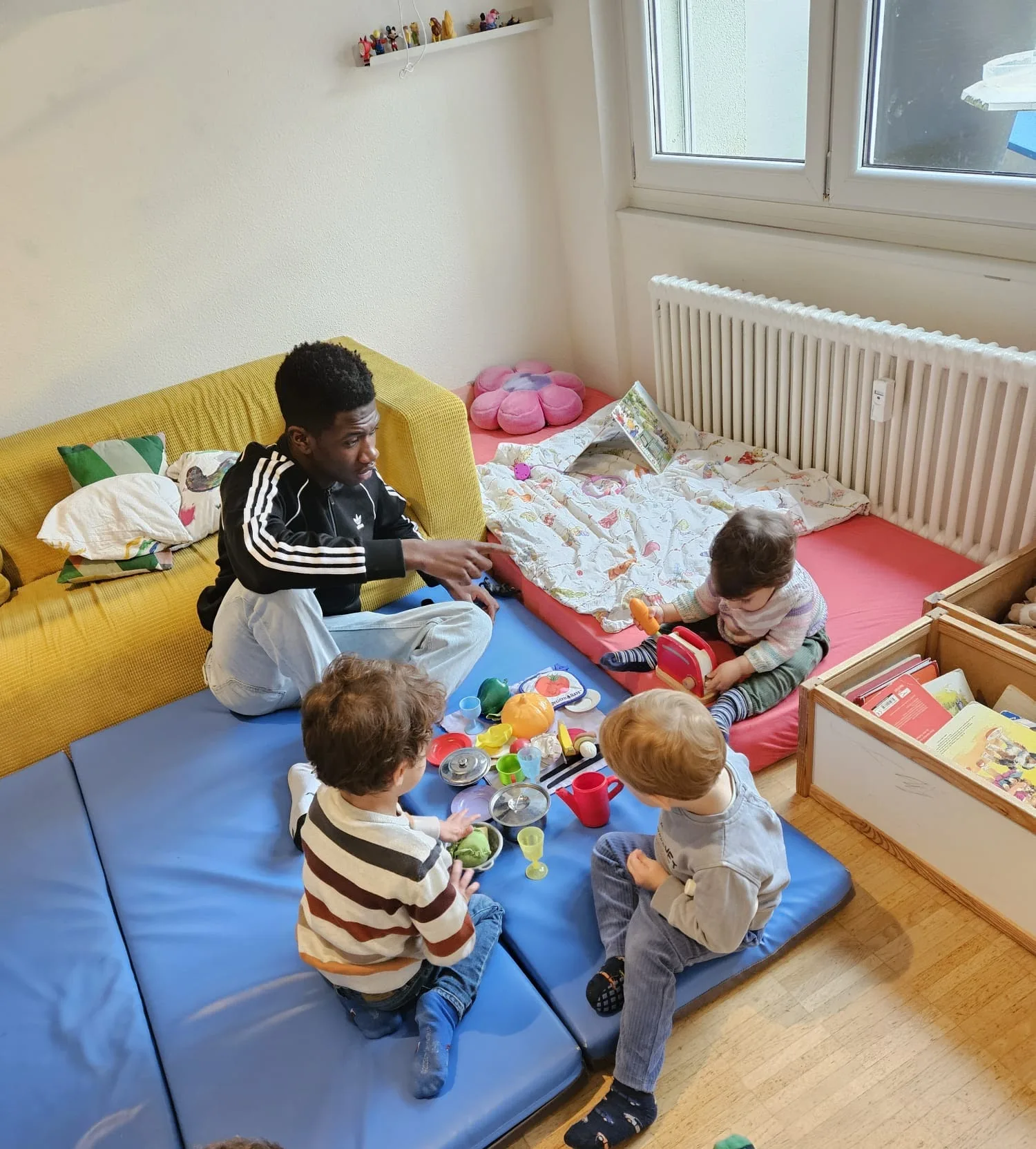 Kinder spielen auf blauen Matten mit Spielzeug im Gruppenraum.