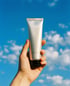 Hand hält eine silberne Tube vor blauem Himmel mit weißen Wolken.