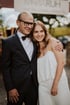 A smiling man in a suit and bow tie with his arm around a young woman in a white dress.