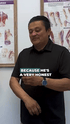 Man in black polo shirt with watch, talking, standing in front of two medical anatomy charts. Text: "BECAUSE HE'S A VERY HONEST".