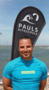 Paul, Gründer und Kitelehrer von Pauls Kiteschule, lächelt vor dem Meer und der Schulflagge am Brouwersdam