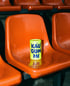 Gelber Becher mit der Aufschrift „KAU GUM MI“ auf einem orangefarbenen Stadionsitz.