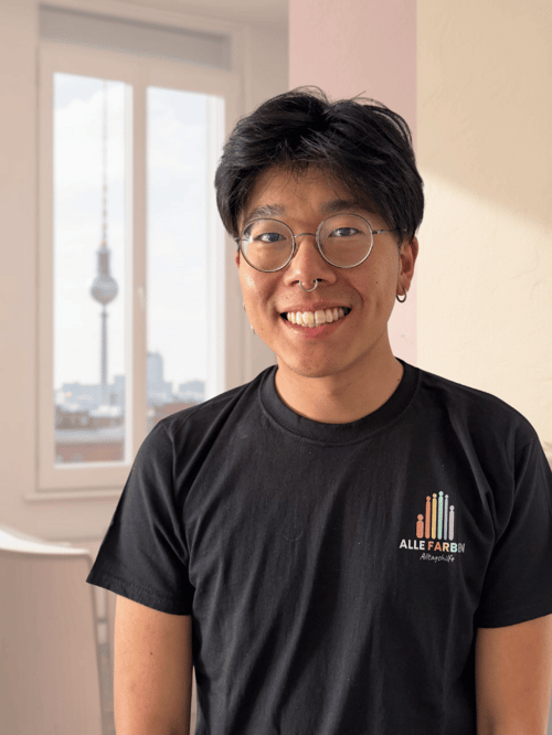 Ein lächelnder Mann mit Brille und Nasenring trägt ein schwarzes T-Shirt mit dem Logo "ALLE FARBEN Alltagshilfe" und steht vor einem Fenster mit Blick auf den Berliner Fernsehturm.