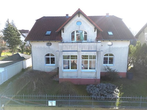 Großes, hellgraues Haus mit rotem Ziegeldach, Balkon, vielen Fenstern und einem Vorgarten.