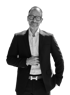 Schwarz-Weiß-Porträt eines lächelnden Mannes mit Brille, Bart, dunklem Sakko und weißem Hemd vor schwarzem Hintergrund.