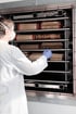 Person in weißem Kittel und Maske legt Handschuh-tragend Bücher in ein Regal in einem Metallschrank.
