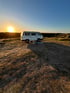 Weißer Campingbus auf sandigem Boden, mit Sonnenuntergang über grünen Dünen und klarem blauem Himmel.