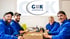 Vier lächelnde Männer in blauen Hemden sitzen an einem Tisch mit einer Pizzaschachtel und G&K-Logo im Hintergrund.