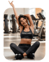 Eine lächelnde Frau in schwarzem Nike Pro Sport-BH und Leggings sitzt kreuzbeinig und dehnt sich in einem Fitnessstudio.