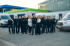 Eine Gruppe von 16 Mitarbeitern in schwarzen Uniformen steht vor einem Gebäude und zwei weißen Lieferwagen.