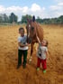 Zwei lachende Kinder stehen neben einem braunen Pferd auf einem Reitplatz im Freien und streicheln es.