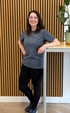 Lächelnende Frau namens Sabrina mit Brille und grauem T-Shirt lehnt an einem Stehtisch vor einer Holzlamellenwand.