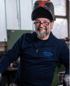 Lächelnder Schweißer mit Schutzhelm, Brille und Bart, trägt einen blauen Pullover mit "BERGMEISTER METALLBAU" Schriftzug.